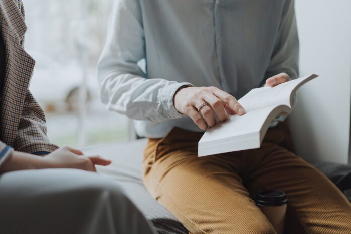 man in white dress shirt and brown pants sitting on white chair reading book