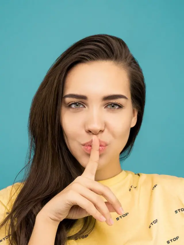 portrait photo of woman in yellow t shirt doing the shh sign while standing in front of blue background